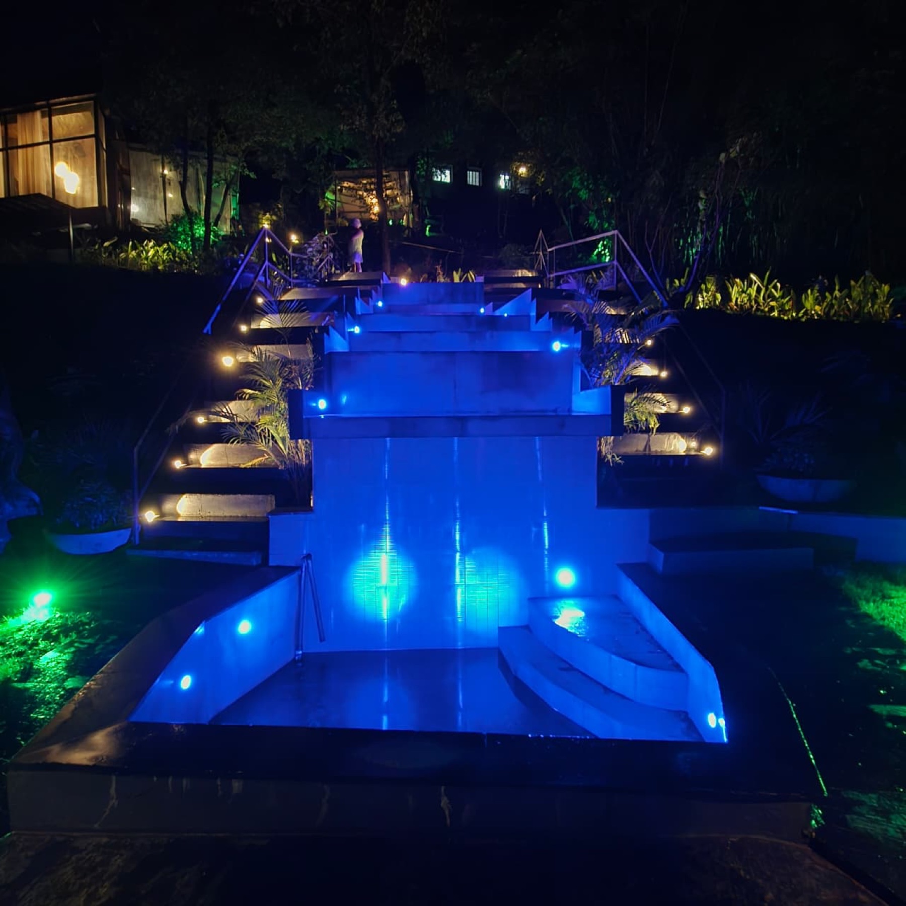 The water cascade lit blue at night with lantern-lit basalt steps rising on both sides